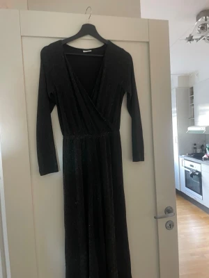 Svart glittrig byxdress - Svart långbyxdress från Kappahl med glittrigt tyg och omlott upptill. Klänningen har markerad midja. I barnstorlek 152