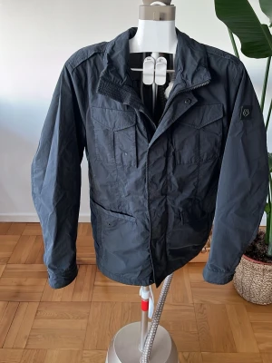 Svart vindjacka från Milton - Svart vindjacka från Milton med flera fickor framtill och diskret logotyp på ärmen. Jackan har dragkedja och knappstängning, samt klassisk krage. Perfekt för dig som gillar en stilren och funktionell look.