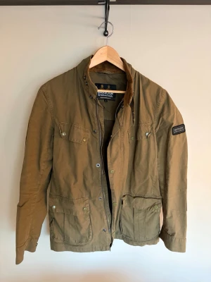 Barbour jacka - Snygg olivgrön jacka från Barbour med klassisk design, flera fickor framtill och brunt manchestertyg på kragen. Jackan har knappar och dragkedja samt patch på ena ärmen. Perfekt för dig som gillar tidlös stil.