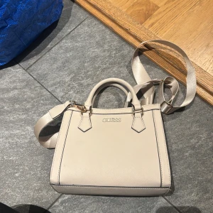 Beige handväska från Guess - Stilren beige handväska från Guess med både handtag och justerbar axelrem. Väskan har en fyrkantig form, guldfärgade metalldetaljer och Guess-logga framtill. Helt oanvänd.