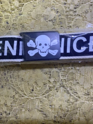Svart bälte med dödskalle och text - Coolt svart bälte med vit dödskalle och korslagda ben på metallspännet. Bältet har även stor vit text längs bandet. Perfekt för dig som vill sticka ut och addera en edgy detalj till din outfit.