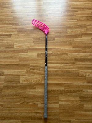 Unihoc  - Säljer en innebandyklubba med svart skaft och ett grått grepp. Bladet är i en stark rosa färg och har en böjd form för bättre bollkontroll. Perfekt för dig som vill sticka ut på planen och gillar sportiga tillbehör.