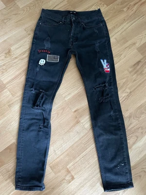 Svarta jeans med patches och slitningar - Säljer ett par svarta skinny jeans med coola patches som amerikansk flagga, smiley, peace-tecken och stjärna. Jeansen har flera slitningar och hål på benen för en trendig look. Perfekta för dig som vill sticka ut lite extra. Materialet är denim.