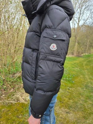 Svart Moncler dunjacka med huva - Svart dunjacka från Moncler med avtagbar huva och klassisk logotyp på ärmen. Jackan har stora quiltade paneler, ficka med knapp på ärmen och dragkedja samt knappar framtill. Perfekt för kalla vinterdagar och riktigt snygg till jeans. Jackan passar som en S/M 
