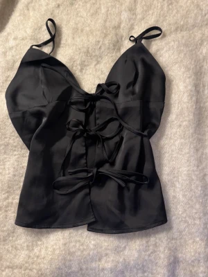 Svart satinlinnetopp med snörning - Snygg svart linnetopp i satin med smala axelband och trendig snörning framtill. Toppen har en lätt glansig finish och en figurnära passform som ger en stilren och modern look.