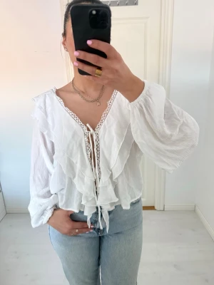 Vit volangblus med knytband - Supersöt vit blus med volanger och knytband framtill. Blusen har v-ringning med spetsdetaljer längs kanten och vida, långa ärmar i ett tunt, luftigt tyg. Perfekt att styla med jeans för en trendig look.