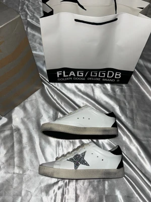 Golden Goose vita sneakers med glitterstjärna - Golden Goose sneakers i vitt läder med svart glitterstjärna på sidan och svart hälparti. Skorna har vita snören, grå sula och klassisk låg profil. Perfekta för dig som vill ha en unik och trendig look med en touch av glitter.24cm 