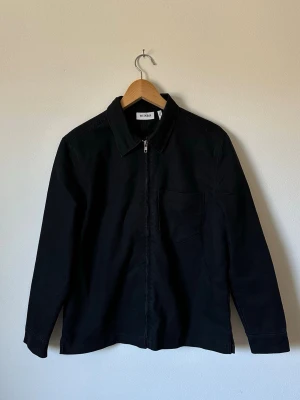 Svart jacka från Weekday - Stilren svart skjortjacka från Weekday med klassisk krage, dragkedja framtill och en bröstficka. Overshirten har raka ärmar och är gjord i ett kraftigt bomullsmaterial som ger en snygg och enkel look. 6R15