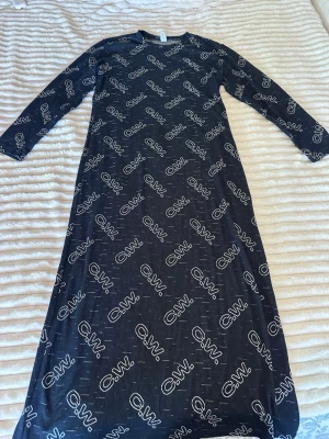 Svart långklänning med logotypmönster - Cool svart långklänning med vitt logotypmönster över hela plagget. Klänningen har lång ärm och rak passform, perfekt för dig som gillar en avslappnad men ändå trendig stil. Tillverkad i mjukt material som känns skönt mot huden.