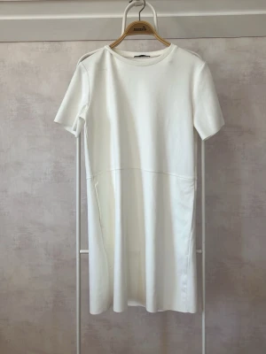 Vit kortärmad klänning - Minimalistisk vit klänning med rund hals och korta ärmar. Klänningen har en rak passform och söm i midjehöjd som ger en stilren look. Perfekt för dig som gillar enkel och clean stil. Materialet känns mjukt och bekvämt.