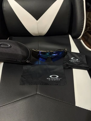 Oakley solglasögon - Snygga sportiga solglasögon från Oakley med spegelblått glas och svart båge. Modellen har en svepande, aerodynamisk form som passar perfekt för utomhusaktiviteter. Kommer med originalfodral och putsduk med Oakley-logga.