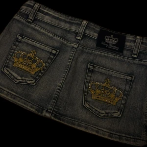 Lågmidjad jeanskjol - Säljer en lågmidjad jeanskjol från Victoria Beckham. Kjolen är blå och har två bakfickor med stora guldbroderade kronor. 