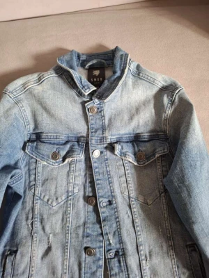 Ljusblå jeansjacka från SMOG - Snygg ljusblå jeansjacka från SMOG med klassisk knappstängning och två bröstfickor med lock. Jackan har en rak passform och coola sömdetaljer som ger en trendig look. Perfekt för dig som gillar streetstyle och denim.