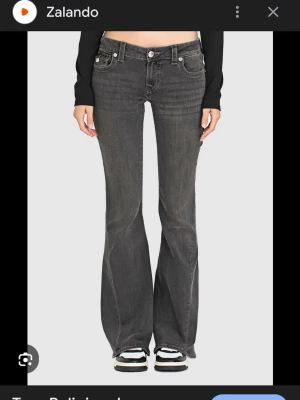 Gråsvarta true religion jeans  - Snygga grå bootcut jeans med låg midja. Jeansen har en klassisk knappgylf och subtila slitningar för en trendig look. Perfekta för dig som gillar en avslappnad men ändå stilsäker vibe. Kan tänka mig att sänka i pris också.