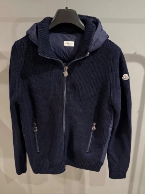 Mörkblå Moncler cardigan tribuet  - Snygg mörkblå jacka från Moncler med stickade ärmar och quiltad kropp. Jackan har huva, dragkedja framtill och två fickor med dragkedja. Moncler-logga på ärmen och ribbade muddar för extra komfort.