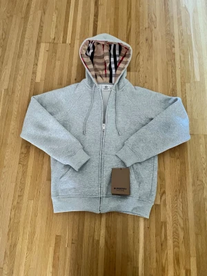 Grå hoodie från Burberry med huva - Snygg grå hoodie från Burberry med dragkedja och klassiskt rutmönster i huvan. Tillverkad i mjuk bomull och har ribbade muddar vid ärmslut och nederkant. Perfekt för dig som vill ha en stilren och exklusiv look. 