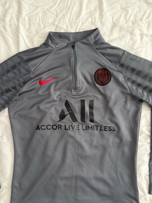 Grå PSG träningströja från Nike - Snygg grå träningströja från Nike med Paris Saint-Germain-logga på bröstet och röd Nike swoosh. Tröjan har halv dragkedja framtill och svarta detaljer på ärmarna. Tillverkad i ett lätt och funktionellt material, perfekt för fotbollsträning.