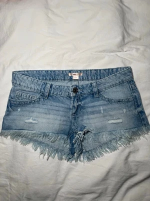 Blå jeansshorts med fransar - Säljer ett par ljusblå jeansshorts från Gina Tricot med fransig kant och slitna detaljer framtill. Shortsen har broderade bakfickor med små pärlor och normal passform. Perfekta för sommaren och riktigt snygga till sneakers eller sandaler.