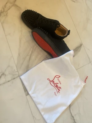 Svarta Louboutin skor med spikar - Säljer ett par svarta sneakers från Christian Louboutin med svarta spikar på tån och klassisk röd sula. Skorna har snörning, låg profil och är tillverkade i mocka med läderdetaljer. Perfekta för dig som vill sticka ut med en edgy och lyxig stil.