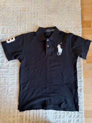Svart pikétröja från Polo Ralph Lauren - Svart pikétröja från Polo Ralph Lauren med klassisk krage och två knappar framtill. Stor vit broderad logotyp på bröstet och siffran 3 på ena ärmen. 