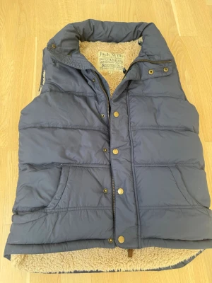 Mörkblå dunväst från Jack Wills - Snygg mörkblå dunväst från Jack Wills med dragkedja och tryckknappar framtill. Västen har två fickor och ett mysigt beige foder i fuskpäls. Perfekt för lager-på-lager och håller dig varm under kyliga dagar.