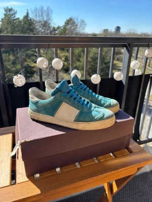 Turkosa sneakers från Valentino - Säljer ett par snygga turkosa sneakers från Valentino med vita detaljer och blå skosnören. Skorna har en låg profil och är tillverkade i mocka och läder. Perfekta för dig som vill sticka ut med färg och stil.