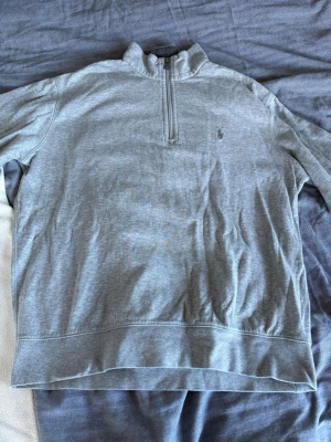 Grå halvzip från Polo Ralph Lauren - Snygg grå stickad tröja från Polo Ralph Lauren med halv dragkedja och hög krage. Tröjan har en broderad logga på bröstet och är perfekt för lager-på-lager. Mjuk bomullsblandning och klassisk passform.