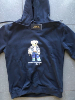 Blå hoodie Polo Ralph Lauren  - Blå hoodie från Polo Ralph Lauren med ikonisk björn-tryck på bröstet. Klassisk huva med dragsko och ribbade muddar. Tillverkad i mjukt material som känns skönt mot huden. Perfekt för dig som gillar streetwear och vill ha en stilren men lekfull look. (Rep)