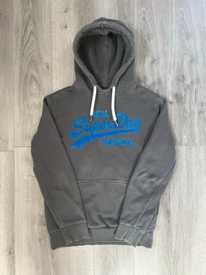Mörkgrå hoodie från Superdry - Säljer en grå hoodie från Superdry med stor blå logga och texten 'Real Superdry Original' på bröstet. Passar Storlek S och inga synliga fel🤩 Priset ej hugget i sten så det är diskuterbart😇