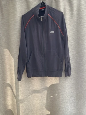 Mörkblå ziptröja från BOSS - Snygg mörkblå ziptröja från BOSS med röda detaljer längs axlarna och vit logga på bröstet. Tröjan har hög krage, hel dragkedja och två fickor framtill. Perfekt för en sportig och avslappnad stil.