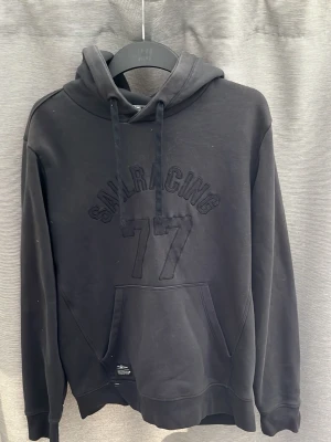 Svart hoodie Sail Racing 77 - Svart hoodie från Sail Racing med broderad text och siffran 77 på bröstet. Klassisk känguruficka framtill, justerbar huva med dragsko och ribbade muddar. Perfekt för en avslappnad och sportig stil.