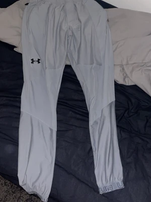 Ljusgrå träningsbyxor Under Armour - Snygga ljusgrå träningsbyxor från Under Armour med elastisk midja och smal passform. Byxorna har dragkedjeficka bak och logga på vänster lår. Tillverkade i ett lätt och stretchigt material som passar perfekt för träning. De finns två prick skador på sista bilden.