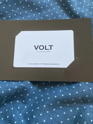 Presentkort från Volt 400 kr - Säljer ett presentkort från Volt, insatt i ett brunt kortfodral. Kortet är vitt med svart text och har Volt-loggan samt texten 'A universe of premium brands'. Perfekt för shopping på voltfashion.com.