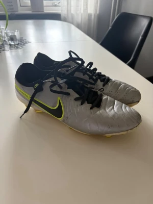 Nike Tiempo Legend silver fotbollsskor - Snygga Nike Tiempo Legend fotbollsskor i silver med svart och neongul swoosh. Skorna har klassisk låg profil, svarta skosnören och dobbar för spel på gräs. Ovandelen är tillverkad i syntetmaterial för bra bollkontroll och komfort. Skriv för mer frågor 