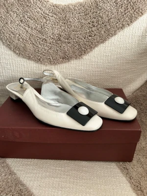 Vita ballerinaskor med svart detalj - Stilrena vita ballerinaskor från  med låg klack och öppen häl. Skorna har en fyrkantig tå och en svart dekorativ detalj med vit knapp framtill. Tillverkade i läder med silverfärgad innersula för en lyxig känsla. Made in Italy. Inköpt i Paris.