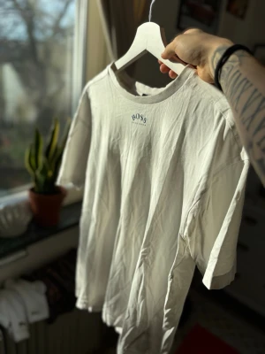 Vit oversized t-shirt från Hugo Boss - Säljer en vit oversized t-shirt från Hugo Boss med liten logga framtill och stor BOSS-tryck på ryggen. T-shirten har rund hals och korta ärmar. Perfekt för dig som gillar stilrena och klassiska plagg med en twist.