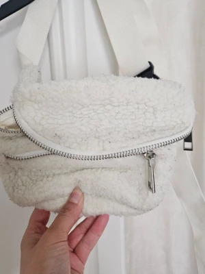 Vit axelväska i teddy-material - Söt axelväska i vitt teddy-material med bred justerbar axelrem. Väskan har ett stort fack med dragkedja i silver och en mjuk, fluffig yta. Perfekt för dig som gillar mysiga accessoarer och vill sticka ut med en unik look.