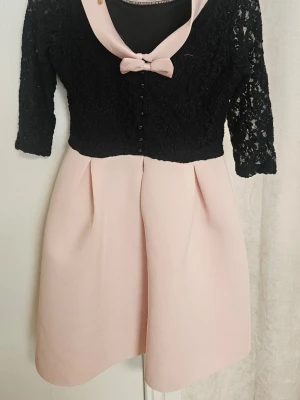 Rosa och svart klänning med spets - Supersöt klänning med svart spetstopp och trekvartsärmar, ljusrosa kjol och matchande rosett vid halsringningen. Kjolen är utsvängd med veck och toppen har små knappar framtill. Perfekt för dig som gillar en feminin och elegant stil.