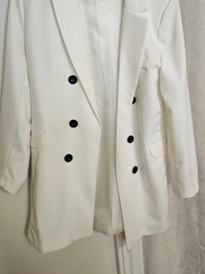 Vit dubbelknäppt kavaj - Stilren vit kavaj med dubbelknäppning och svarta knappar. Klassisk passform med två fickor framtill och breda slag. Perfekt för en trendig och clean look. Materialet ser ut att vara en slät och lätt tygblandning.