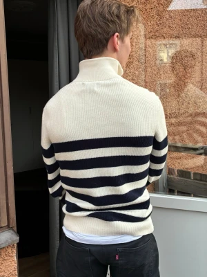 Randig stickad tröja med halvzip - Snygg stickad tröja med breda marinblå och vita ränder, halvzip i halsen och ribbade muddar. Perfekt för dig som gillar klassisk men trendig stil. Tröjan har hög krage och är gjord i ett mjukt, tjockt material som håller värmen.