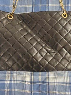 Svart quiltad handväska med guldkedja - Snygg svart handväska i quiltat mönster med guldfärgade kedjehandtag och detaljer. Väskan har en rektangulär form och är tillverkad i ett glansigt material som liknar konstläder. Perfekt accessoar för att lyfta din outfit.