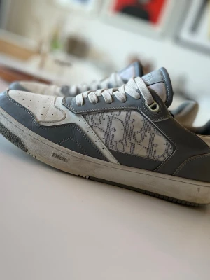 Dior gråvita sneakers med monogram - Snygga Dior sneakers i grått och vitt med klassiskt Dior-monogram på sidan. Skorna har lågt skaft, perforerad tå och vita skosnören. Tillverkade i läder och textil med robust gummisula där Dior-loggan syns tydligt.