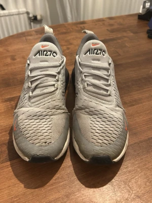 Nike Air Max 270 grå sneakers - Nike Air Max 270 sneakers i grått med orange detaljer och svart sula. Ovandelen är i mesh för bra andningsförmåga och skon har en tydlig Air Max-dämpning i hälen. Snygg och sportig design med klassisk Nike-logga på sidan och tungan.