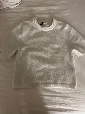 Vit stickad topp från H&M Divided - Mysig vit stickad topp från H&M Divided med korta ärmar och ribbade detaljer vid hals, ärmslut och nederkant. Perfekt till jeans eller kjol för en enkel och clean look.