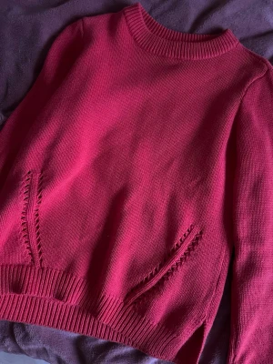 Mörkrosa stickad tröja - Säljer en mörkrosa stickad tröja med rund halsringning och ribbade muddar vid ärmslut och nederkant. Tröjan har dekorativa stickade detaljer vid fickorna framtill och är i mjukt material. Perfekt för dig som gillar färg och enkel stil.