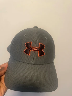 Grå keps från Under Armour - Snygg grå keps från Under Armour med det klassiska logotypen broderad i svart och orange framtill. Kepsen har böjd skärm och är tillverkad i ett ventilerande, strukturerat material som ger en sportig look.