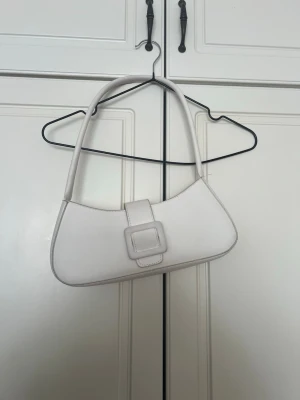 Vit axelremsväska med spänne - Snygg vit handväska i minimalistisk design med axelrem och dekorativt fyrkantigt spänne framtill. Väskan har en avlång, böjd form och är tillverkad i ett slätt material som liknar skinn. Perfekt för dig som gillar stilrena accessoarer.se fler väskor på min sida 🤍💕