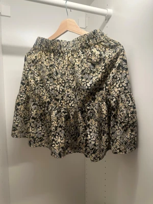 Svart blommig volangkjol - Supersöt svart kjol med vitt blommönster och volangkant. Kjolen har en avslappnad passform med resår i midjan och är perfekt för dig som gillar en feminin och trendig stil. Materialet känns lätt och luftigt.