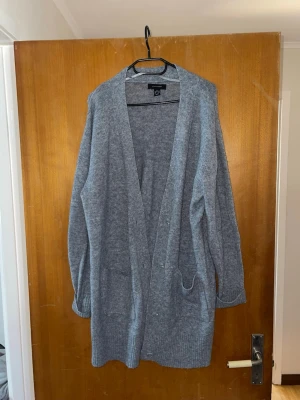 Grå lång kofta från Atmosphere - Mysig grå lång kofta från Atmosphere med öppen front och två fickor. Koftan har ribbade muddar vid ärmslut och nederkant, perfekt för lager på lager. Tillverkad i mjukt stickat material som känns skönt mot huden.