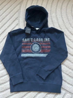 Mörkblå Saint Laurent hoodie - Snygg mörkblå hoodie från Saint Laurent med tryckt logga och emblem på bröstet. Hoodien har dragsko i huvan och en stor magficka. Tillverkad i mjuk bomull för skön känsla och avslappnad passform. Perfekt för dig som gillar streetwear och exklusiva märken. Skickas samma dag, skriv gärna vid frågor😊